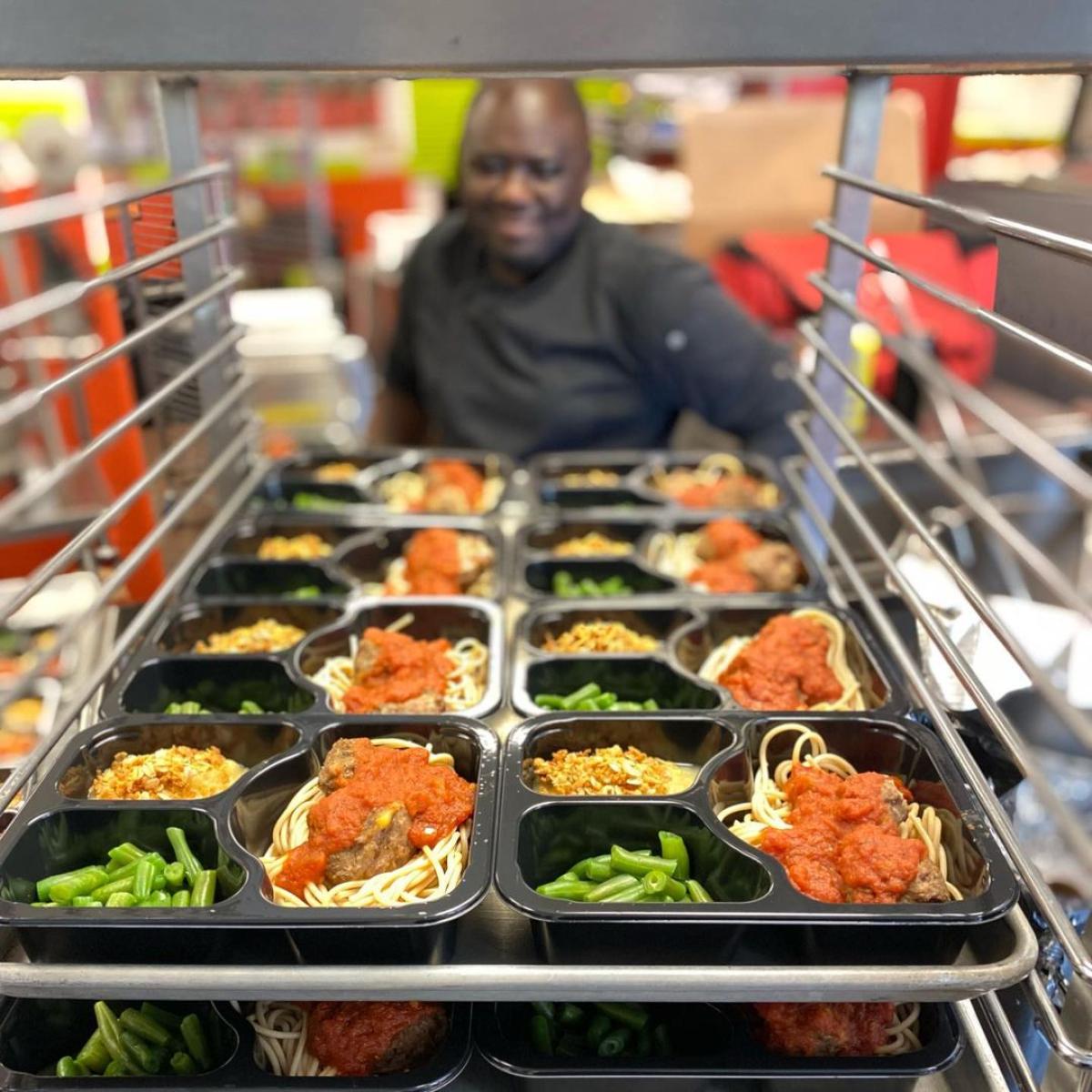 A cart is stacked full of trays of school meals - spaghetti and meatballs, green beans, and apple sauce. A chef smiles in the background.