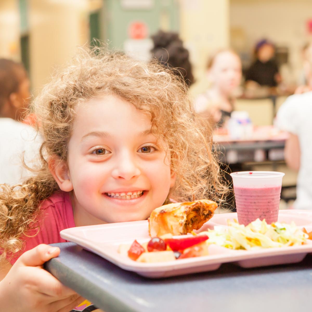 A child smiles, a lunch tray sitting on the cafeteria table in front of them.