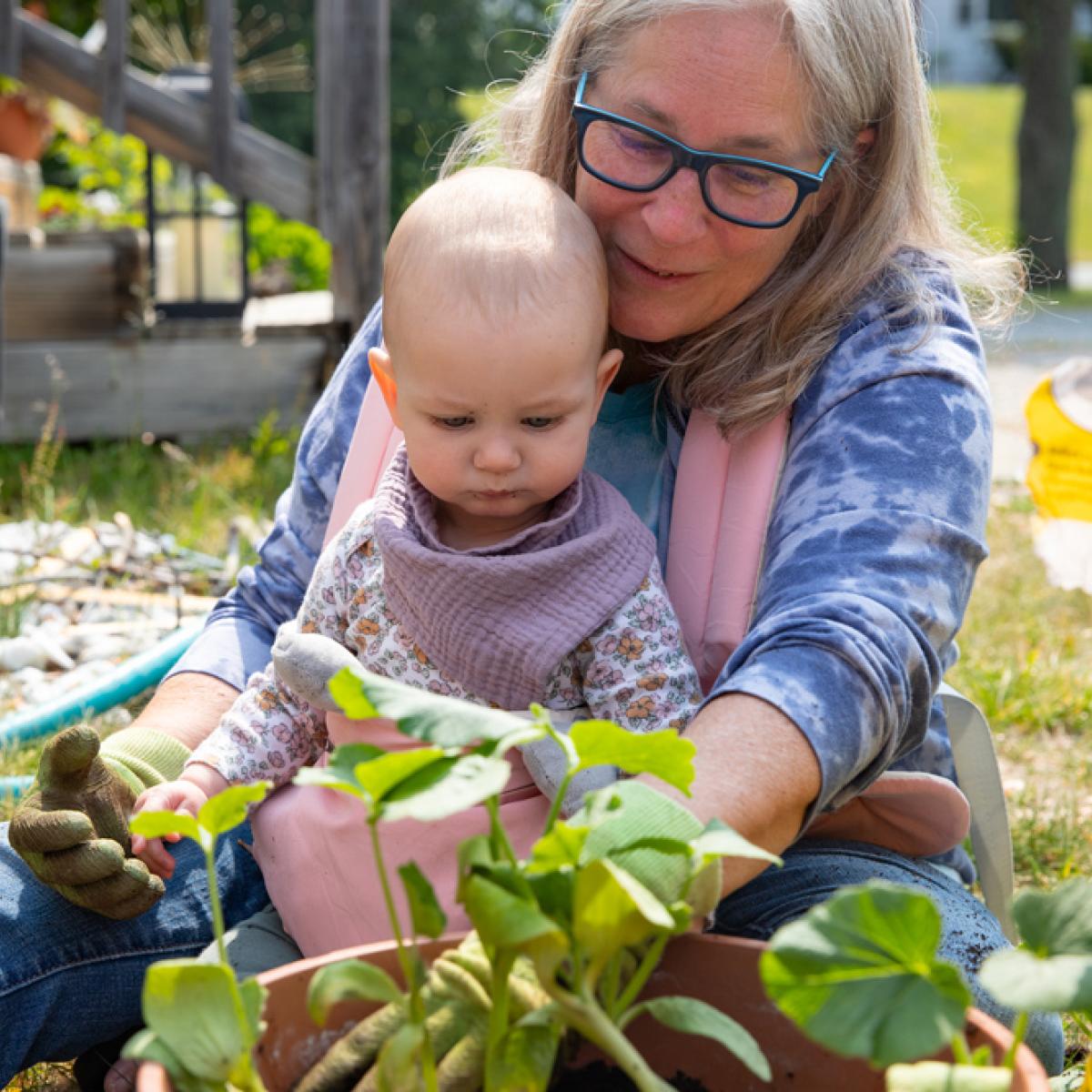 A woman sits with a baby in a carrier attached to her body, they look at a squash plant
