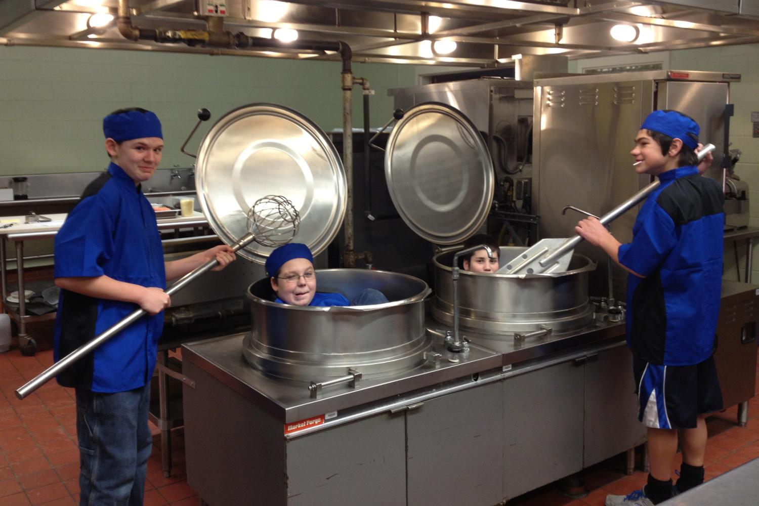 Four boys in a professional kitchen. Two are sitting in large soup pots.