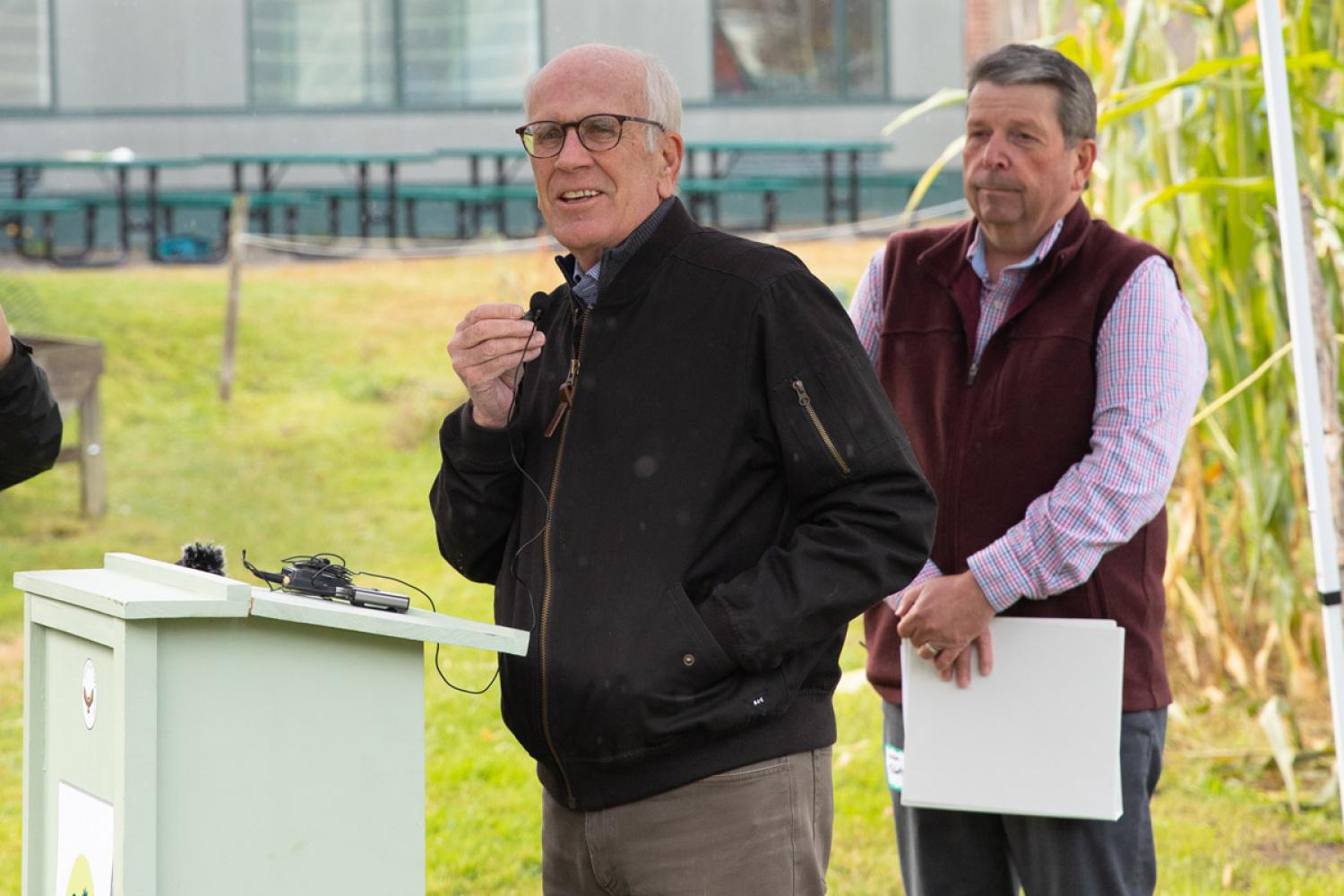 U.S. Senator Peter Welch and Vermont Secretary of Agriculture, Food and Markets Anson Tebbetts stand at an outdoor podium; one is speaking with a microphone and the other is holding a clipboard, with a building in the background.