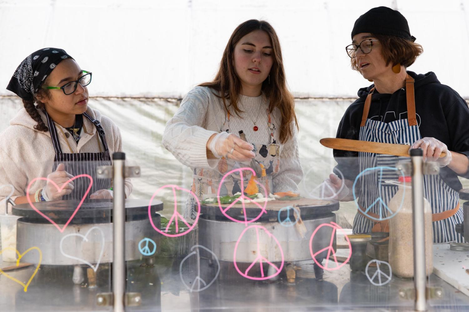 Three people are making crepes in an outdoor cooking cart.