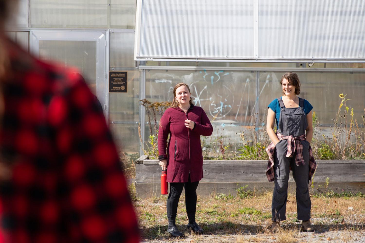 Two individuals stand in front of a greenhouse, one holding a clipboard and the other with hands clasped, smiling and engaging in conversation.