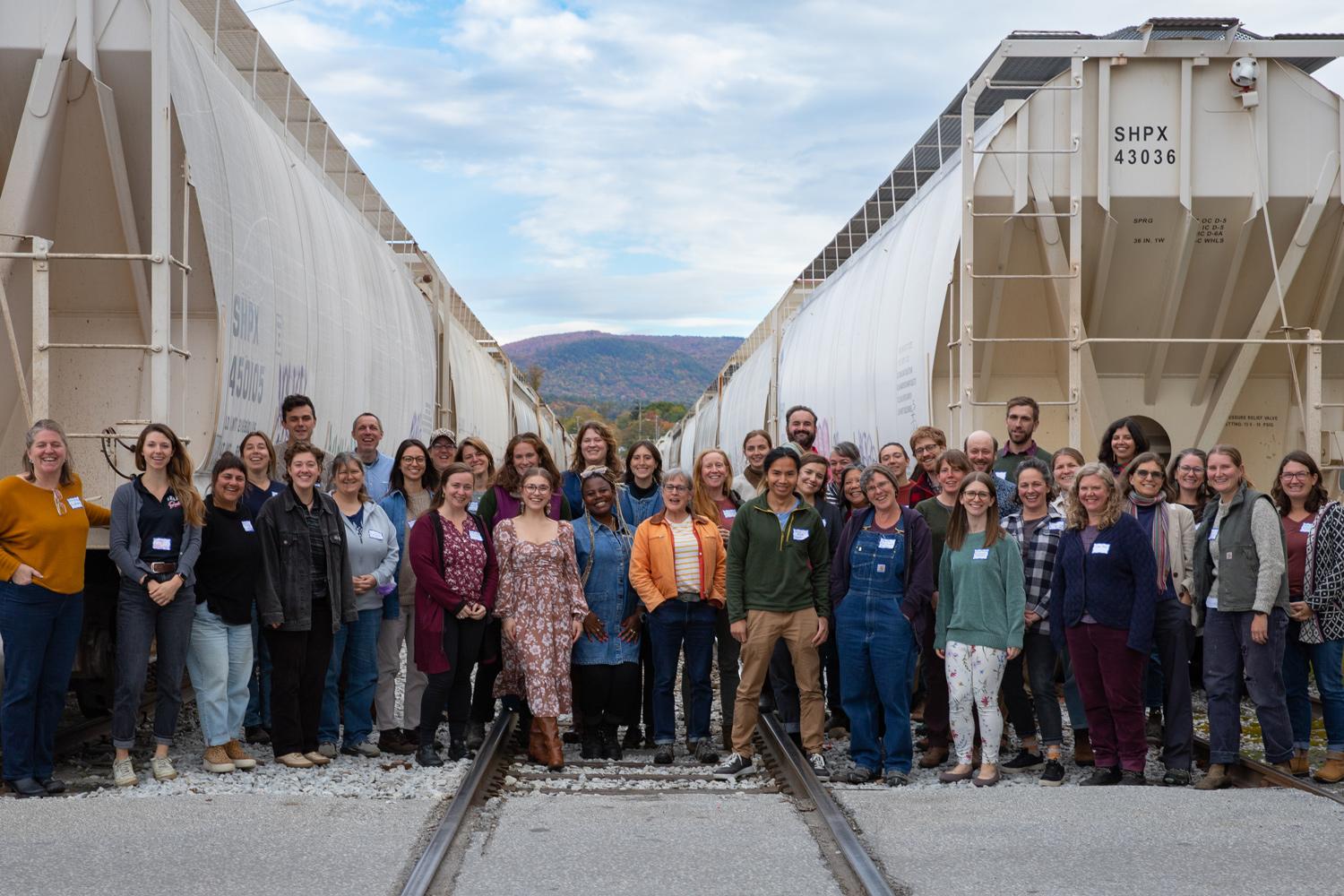 Group of people standing between two rows of white train cars on railroad tracks, with a scenic mountain backdrop.