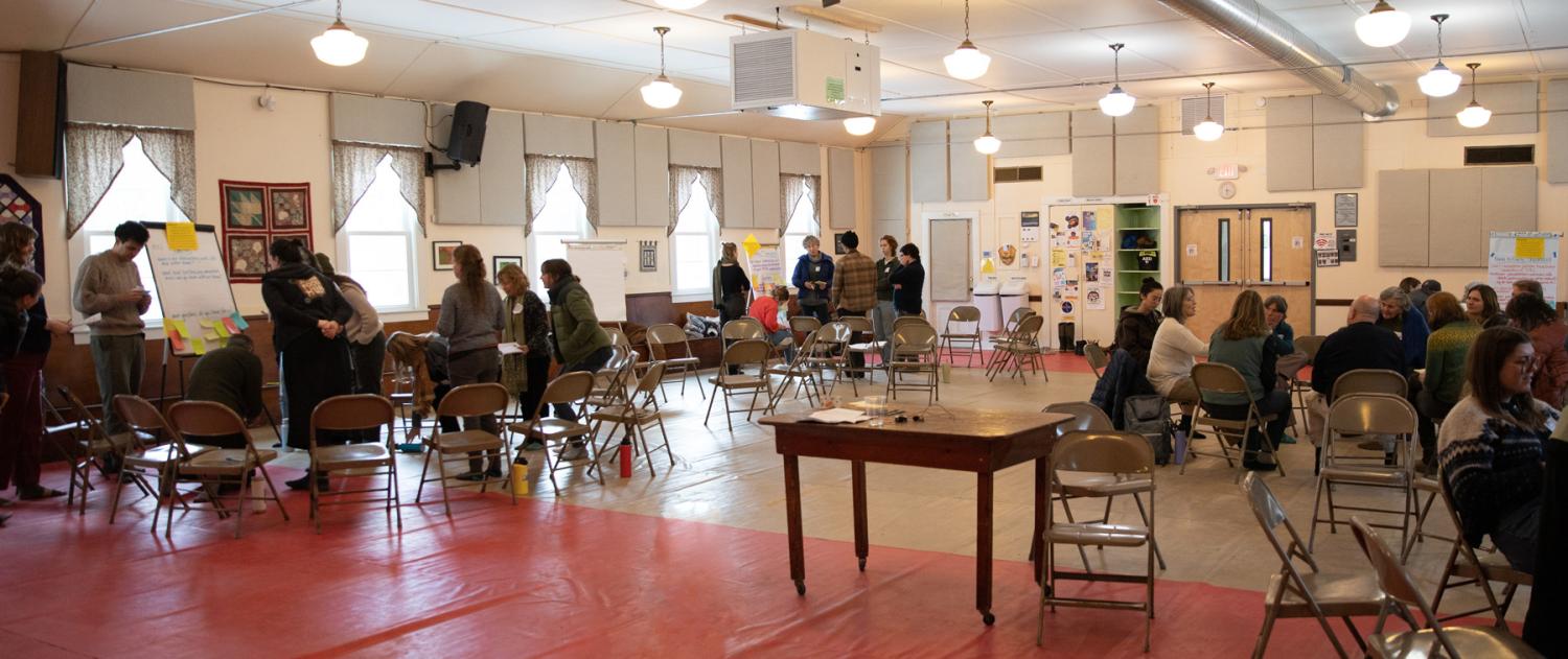 A community center room filled with people seated on folding chairs and engaged in conversation.