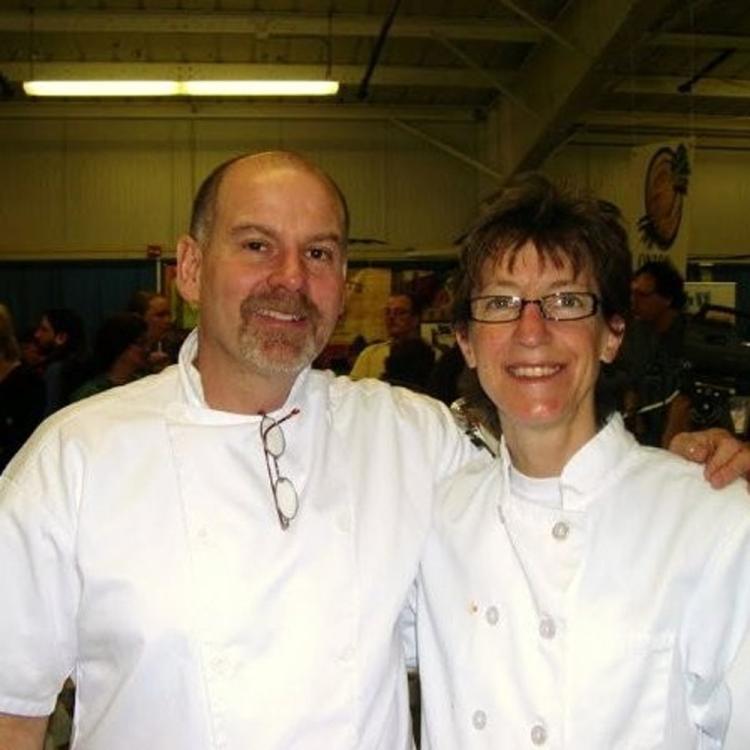 Two chefs in white uniforms smiling at the camera in a busy event setting.