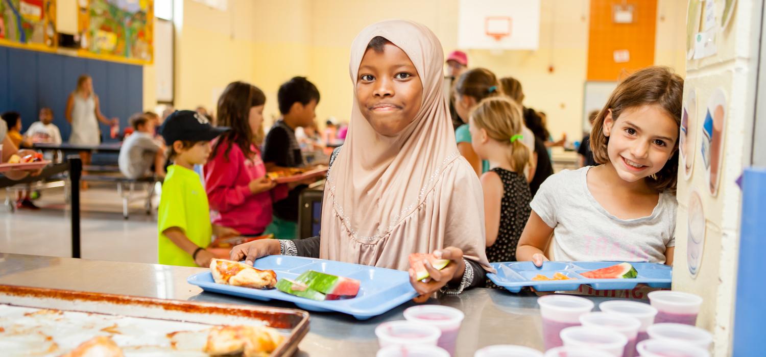 Two children, one wearing a hijab, are selecting food in a school cafeteria, with peers eating at tables in the background.