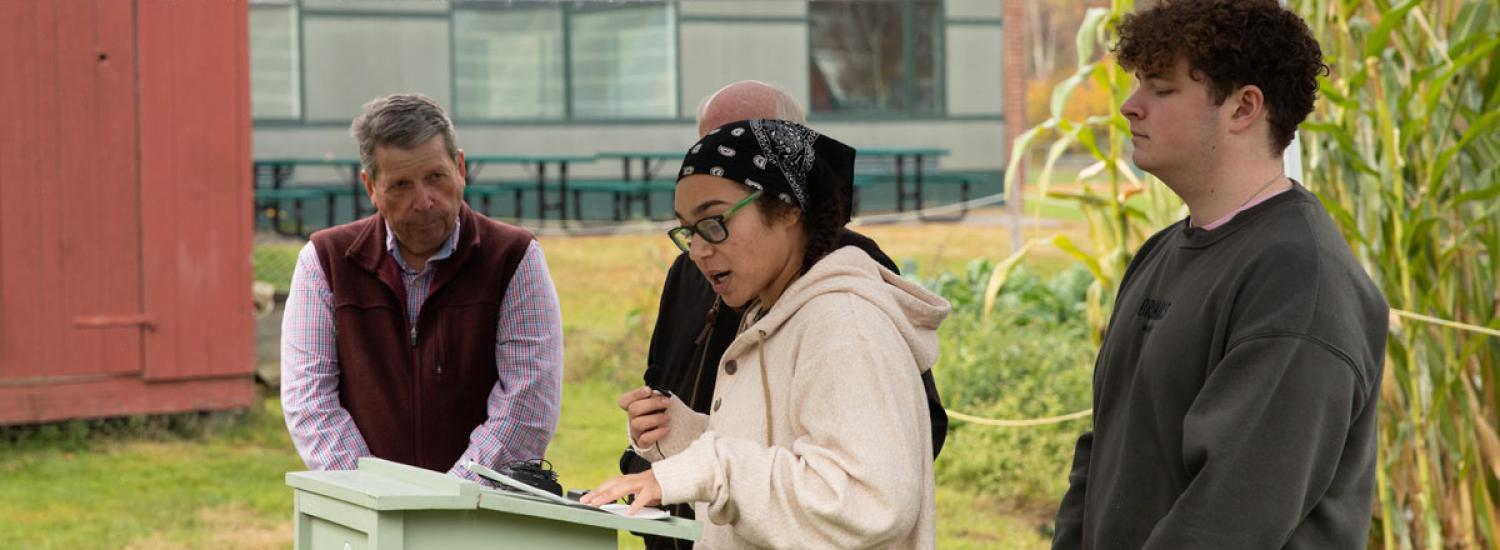 Three people under a tent, one speaking at a podium while the others listen, with greenery and a red building in the background.