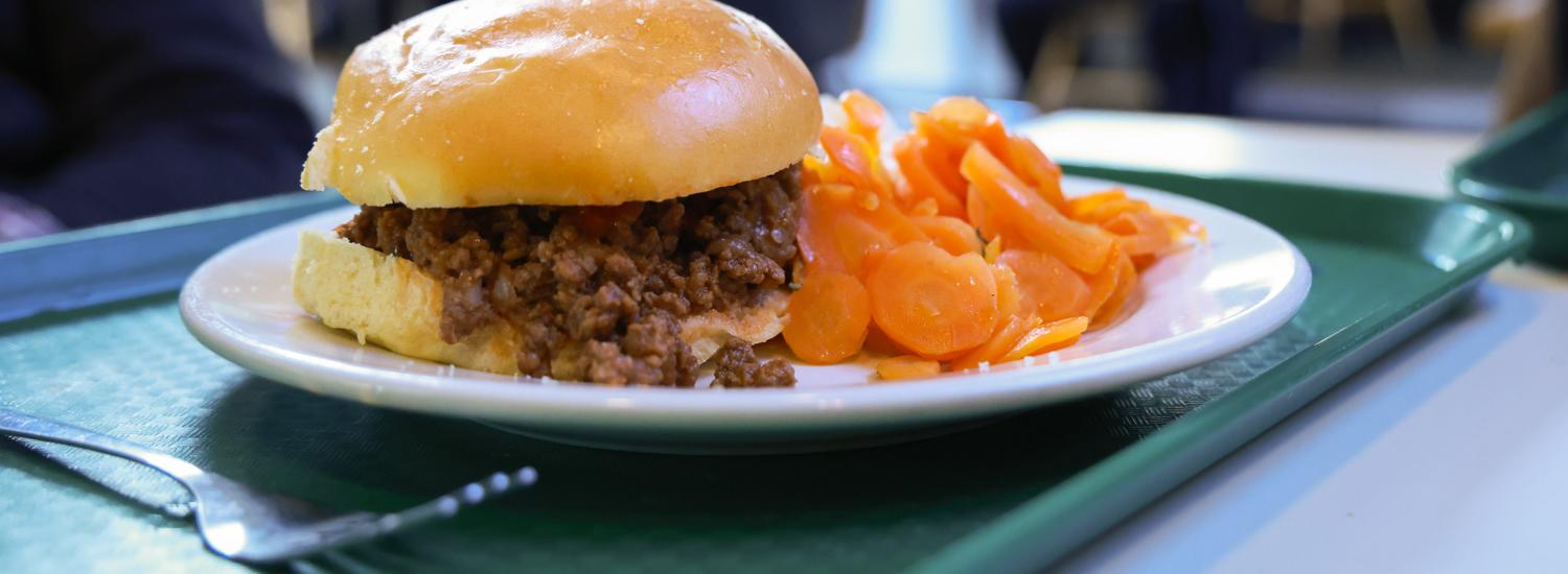 A close-up of a sloppy joe sandwich and a side of sliced carrots on a tray, set on a table in a cafeteria with people in the background.