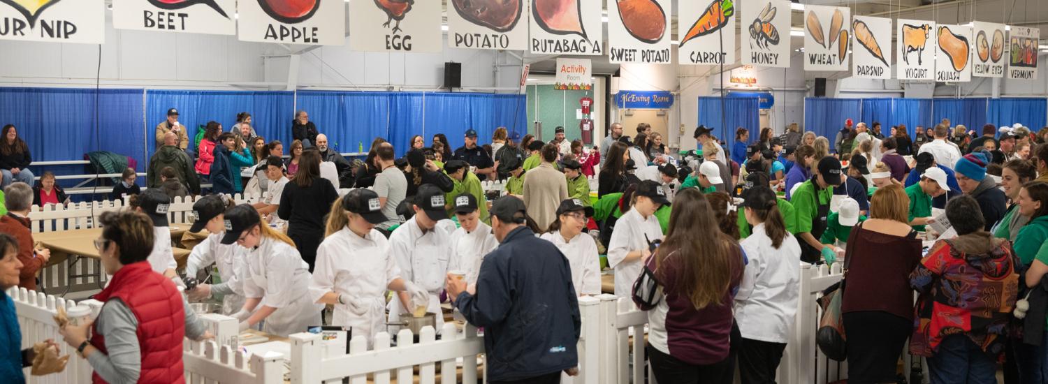 Youth chefs at a bustling culinary competition with various fruit and vegetable banners like Apple, Potato, and Carrot displayed above booths where individuals wearing white chefs' hats are cooking.