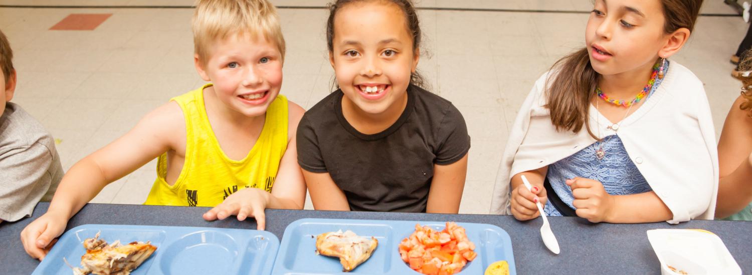 Children smiling at a lunch table with trays of food in a school cafeteria.